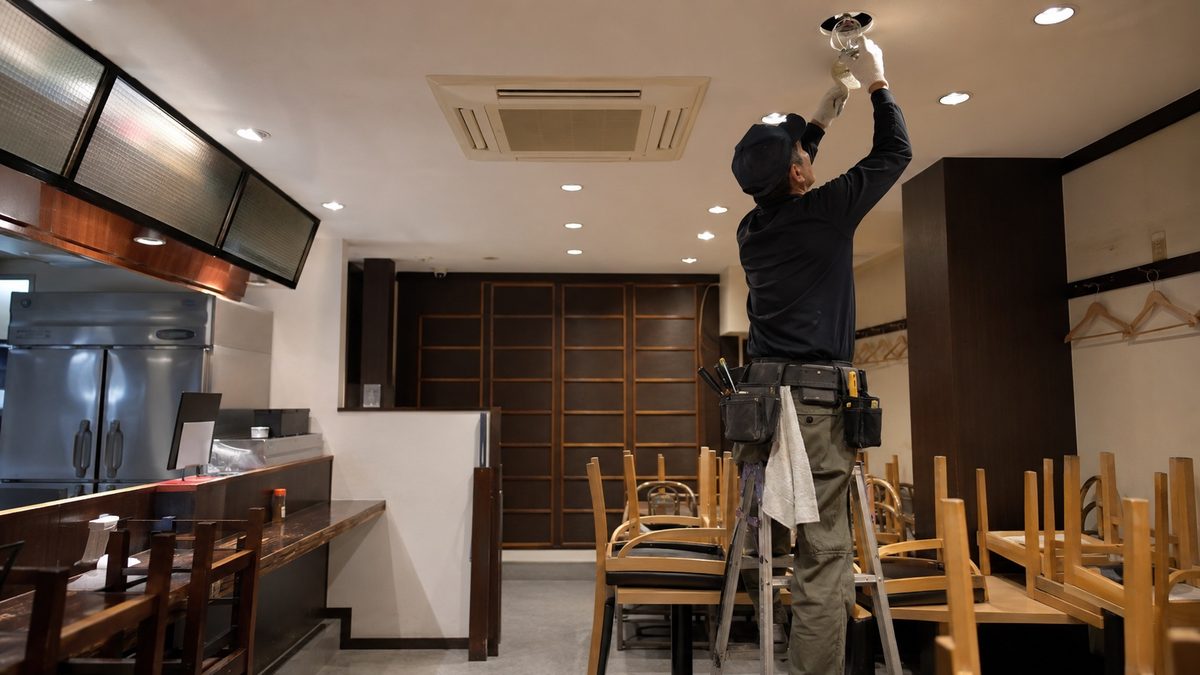 小さな店舗で脚立に立ち天井照明を扱う職人(AI・構成見本)
